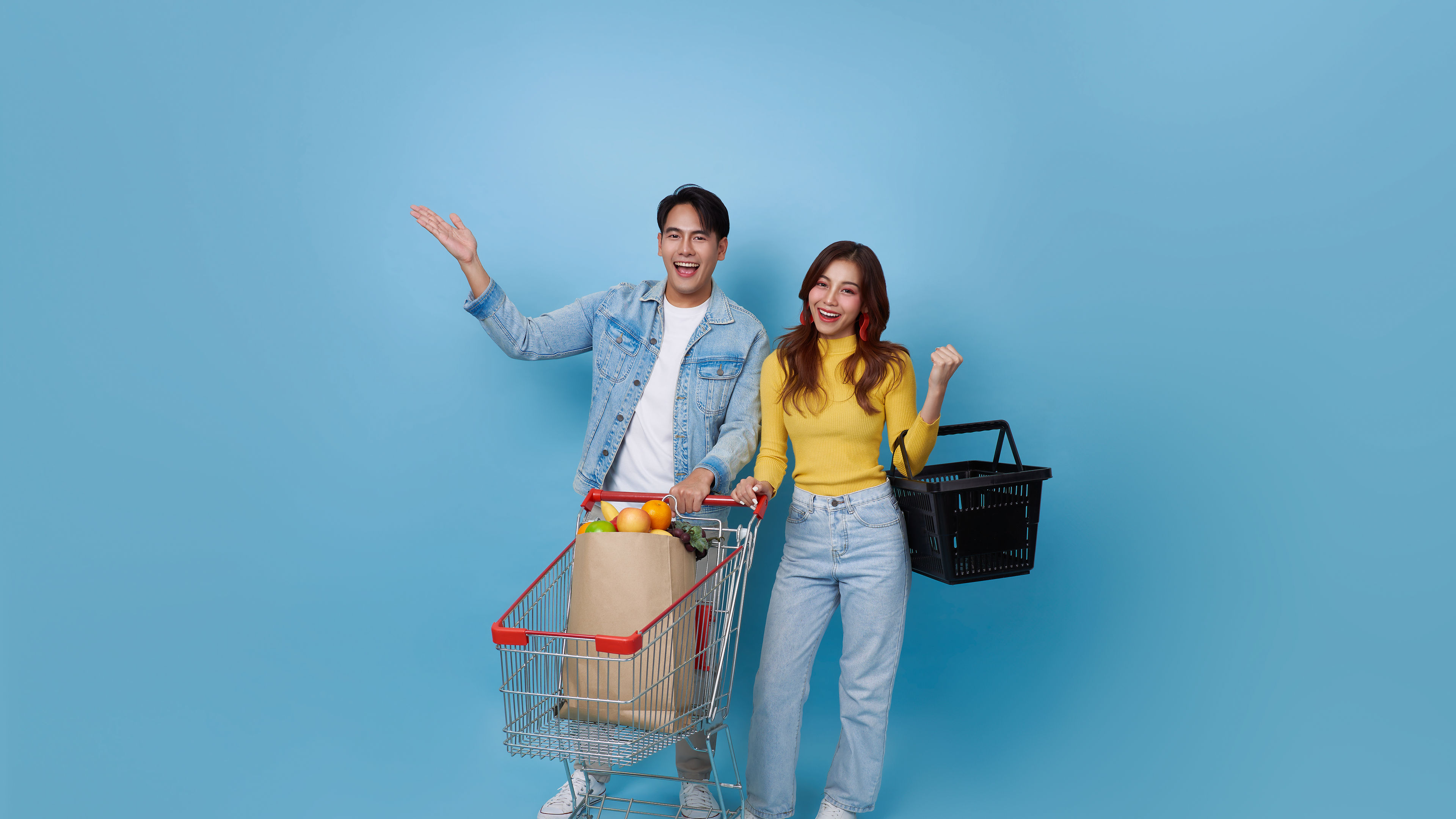 Bata masayahin Asyano mag asawa na may shopping trolley cart at may hawak na shopping bag sa isang asul na background

