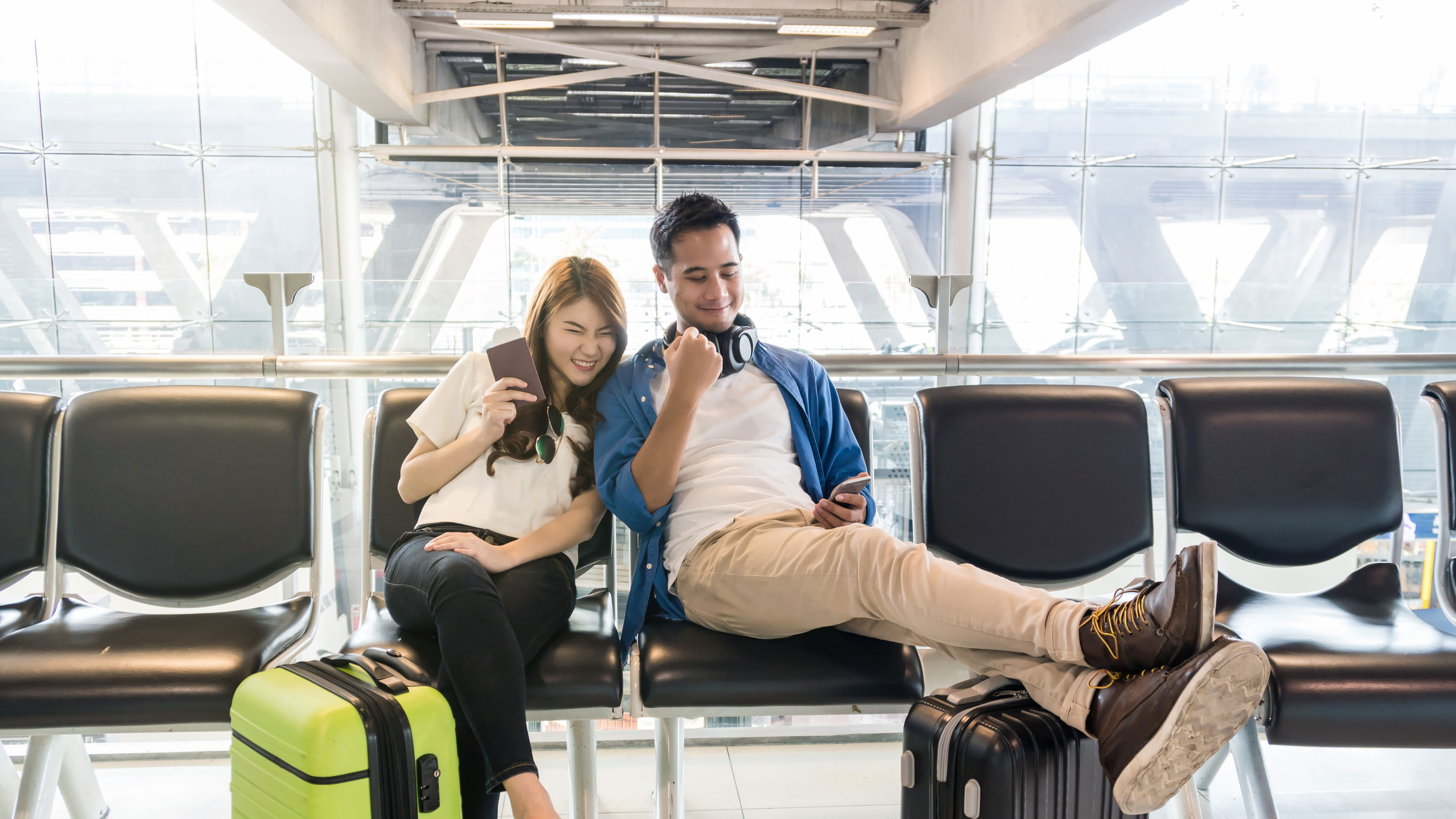 Isang batang mag asawa na nakaupo sa isang terminal ng paliparan, nakangiti at nakakarelaks, napapaligiran ng mga bagahe, na may modernong background na salamin.