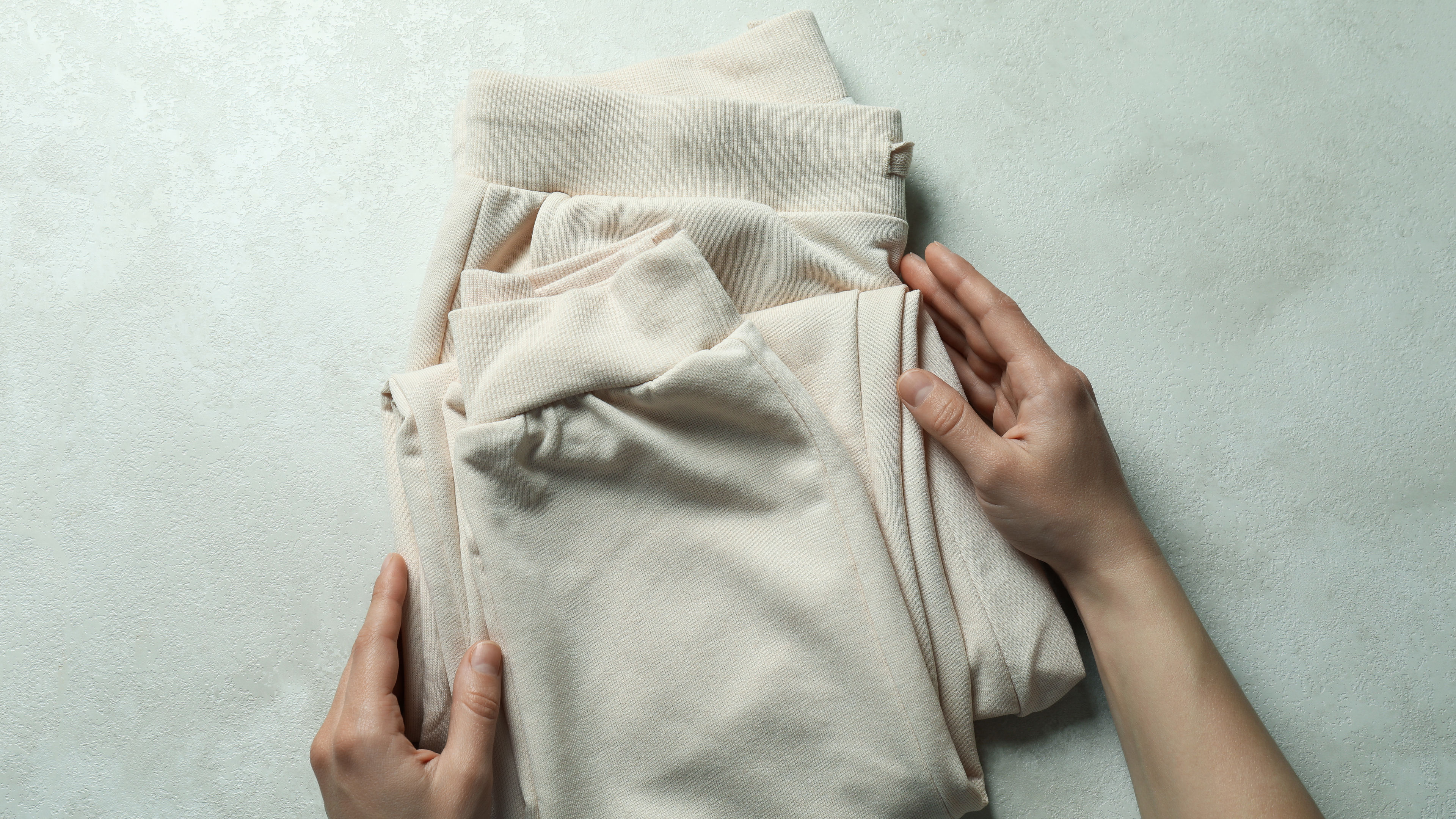 Ang isang pares ng mga kamay ay malumanay na may hawak na nakatiklop na cream-colored sweatpants.
