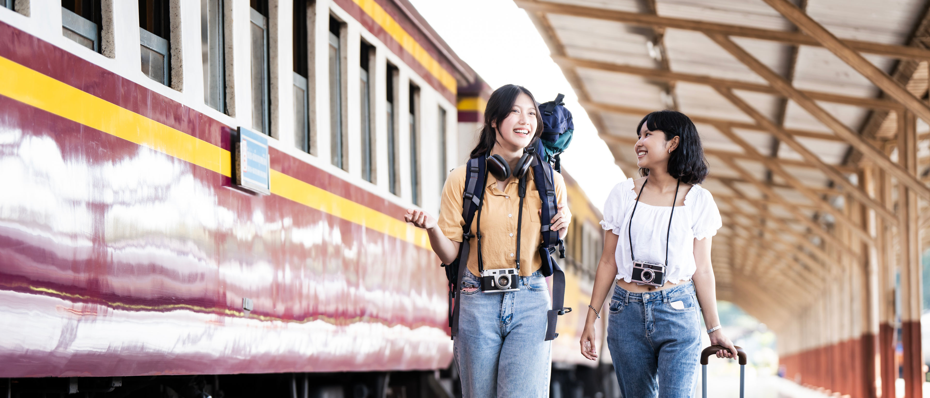 Dalawang kabataang babaeng Asyano na may backpack na magkasamang naglalakad sa Train Station Platform