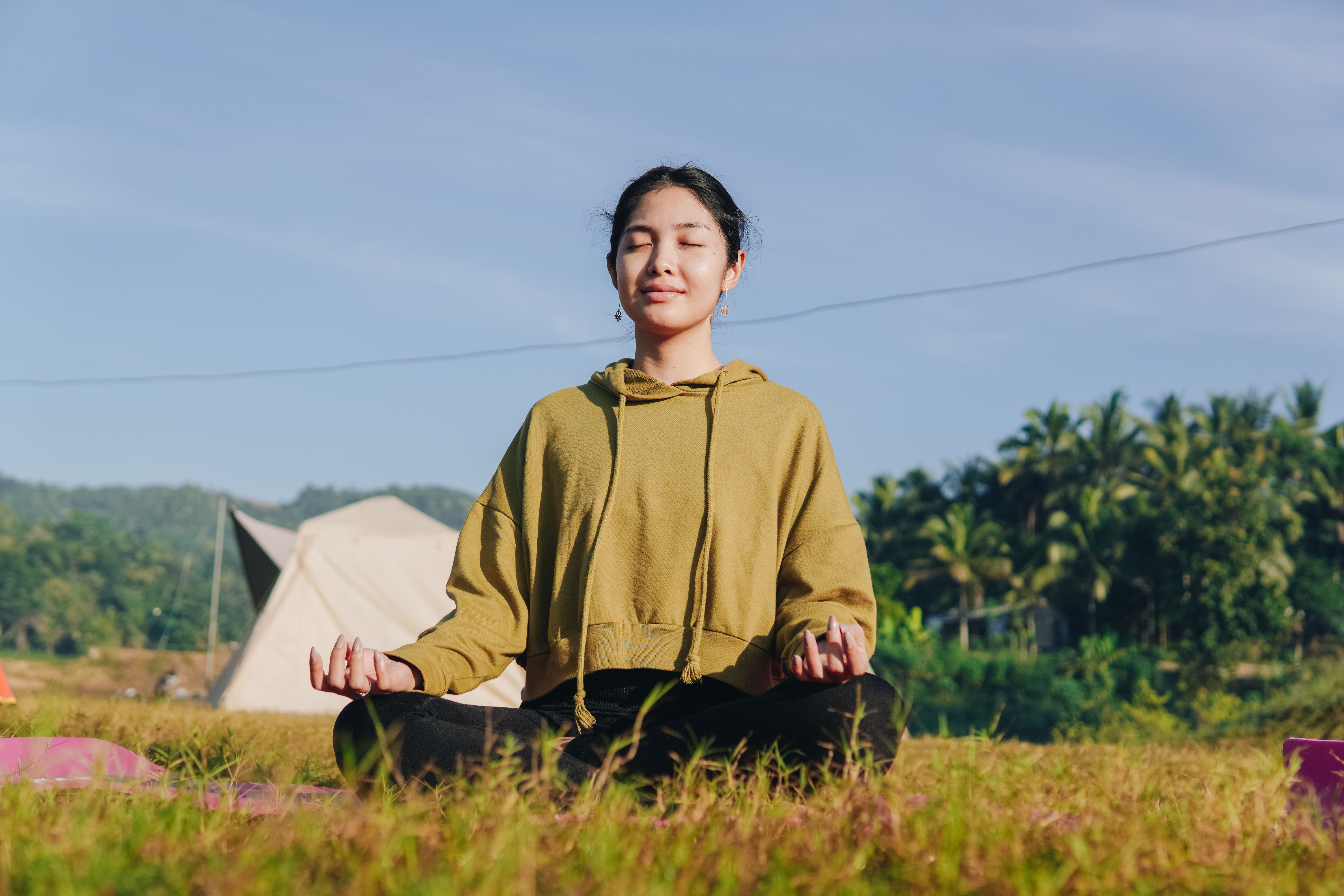 Babae ay nakaupo sa isang patlang, suot ng isang berdeng hoodie at paggawa ng yoga